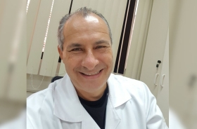 Homem posa para foto sorrindo. Ele está de jaleco em um consultório.