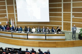 Cerimônia formal em auditório. Autoridades sentadas à mesa no palco, homem discursa no púlpito. Público assiste. Telão ao fundo exibe participante remoto. Bandeiras à esquerda, planta decorativa à direita.