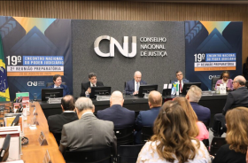 Quatro homens e uma mulher estão sentados em uma mesa de uma palestra. Pessoas estão assistindo. No fundo do palco tem um painel com o texto: CNJ