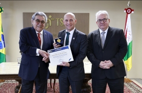 Três homens de terno e gravata estão em pé, posando para a foto. O homem que estão no centro segura um certificado e uma medalha