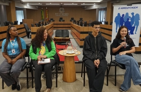 Três jovens e uma mulher madura sentados em cadeiras, dentro de uma ampla sala de julgamentos. Uma das jovens segura o microfone e sorri
