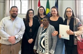 Cinco mulheres e um homem de pé, lado a lado, posam para a foto. Uma das mulheres segura um documento.