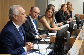 Foto de uma mesa de honra de um evento, com um homem falando e mais homens e mulheres sentados olhando o palestrante