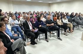 Foto em diagonal mostra cerca de 90 pessoas sentadas em cadeiras dentro de um auditório