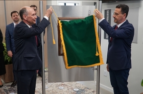 Um homem calvo, de cerca de 60 anos, de terno, e outro homem moreno, também de terno, retirar um pano verde que está cobrindo uma placa de metal