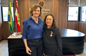 Duas mulheres, uma loira e outra morena, posam sorridentes para a foto. A morena tem uma medalha fixada na roupa