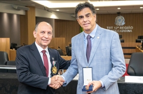Dois homens de terno, com cerca de 60 anos, se cuprimentam. Um deles, de terno claro, exibe uma caixa contendo uma medalha.