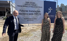 Um homem de terno e duas mulheres ao lado de uma placa de cerca de 2 metros de altura sobre um terreno vazio