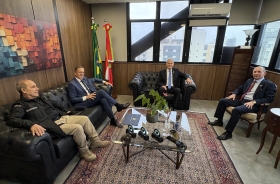 Três homens de terno e um com uniforme de segurança sentados em poltronas e cadeiras em uma sala arejada