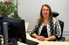 Mulher de pele branca e cabelos castanhos claros, sentada em um posto de trabalho, sorri para a câmera