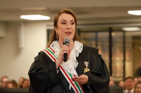 Mulher de toga e faixa fala ao microfone