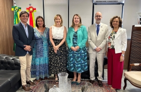 Quatro mulheres e dois homens, todos de pé, posam para a foto em uma pequena sala com bandeiras do Brasil e de SC ao fundo