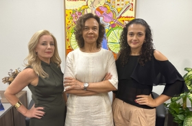 Três mulheres vestidas formalmente em pé, em pose desafiadora, diante de um quadro colorido