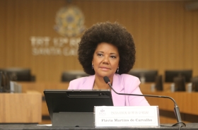 Mulher negra, de cabelo crespo, estilo "black power", fala ao microfone sentada em uma mesa de evento