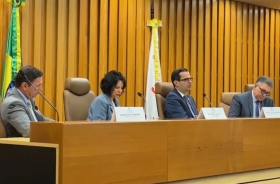 Três homens de terno e uma mulher vestida formalmente sentados em uma mesa de eventos, em tom madeira-ouro, com uma bandeira do Brasil atrás