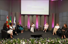 Grupo de sete mulheres sentadas à frente de um auditório, sobre um tablado, formando um semicírculo. Uma delas fala ao microfone