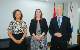 Duas mulheres vestidas formalmente e um homem de terno escuro posam para foto, de pé. Eles estão em uma sala