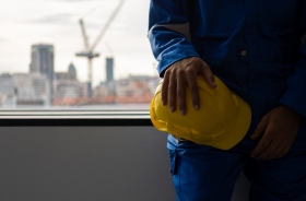 Trabalhador de obra com uniforme azul segura capacete amarelo diante de janela com cidade ao fundo