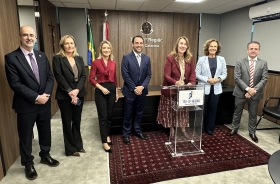 Três homens e quatro mulheres em pé, em uma sala, lado a lado, todos sorridentes. Uma das mulheres assina um documento em um púlpito transparente