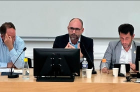 Homem calvo, de óculos, sentado à frente de um notebook, faz uma apresentação. Ao lado dele estão outros dois homens.