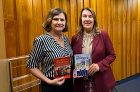 Duas mulheres posam para a foto. Cada uma exibe uma cartilha