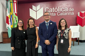 Três mulheres vestidas formalmente e um homem de terno azul marinho posam para a foto, sorridentes, em um auditório. Ao fundo, a logomarca da Universidade Católica