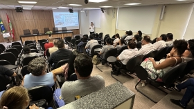Foto de uma palestra, com pessoas sentadas em um auditório a uma mulher palestrante na frente do público 