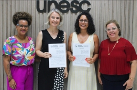 Quatro mulheres estão em pé, posando e sorrindo para a foto, segurando um contrato