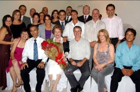 Foto posada de 21 pessoas, algumas sentadas e outras em pé. No centro, Ione Ramos está sentada e segurando, sorridente, uma buque de flores