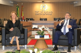 Home de terno escuro fala ao microfone sentado em uma poltrona, na sala de sessões do Pleno do TRT-SC. Ao lado dele, em outra poltrona, uma mulher loira sorridente, com roupa preta