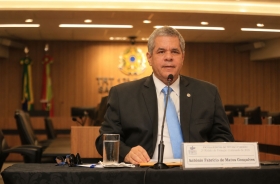 Fotografia de homem sentado falando em uma mesa de honra, durante uma palestra