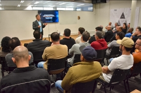 Homem de terno palestrando para grupo de pessoas em um pequeno auditório