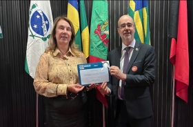 Uma mulher vestida formalmente e um homem de terno e gravata seguram, juntos, um certificado. Ao fundo bandeiras do Brasil e dos estados do Paraná, Santa Catarina e outra não identificada