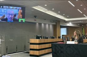 Mulher sentada à mesa de um evento observa palestrante que aparece em um telão