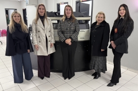 Cinco mulheres posam para a foto. Atrás delas, dois equipamentos de informática