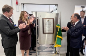 Dois homens de terno e gravata e uma mulher estão aplaudindo felizes em frente a uma placa de inaguração de um forum trabalhista