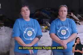 Duas mulheres brancas, uma de cabelos escuros e outra loira, estão lado a lado, olhando para a câmera, à frente de resíduos para reciclagem