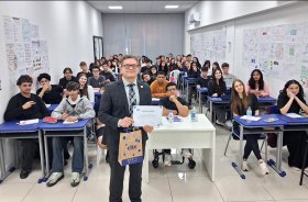 Homem de terno mostrando um certificado em uma sala de aula com diversos adolescentes sentados em carteiras escolares ao fundo
