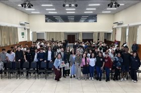 Cerca de cem jovens e adolescentes e outros quatro adultos posam para foto, de pé, em um amplo auditório