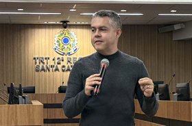 Homem de blusa preta e calça caqui, de pé, fala ao microfone. Ele está em um auditório, tendo ao fundo um brasão do Poder Judiciário iluminado