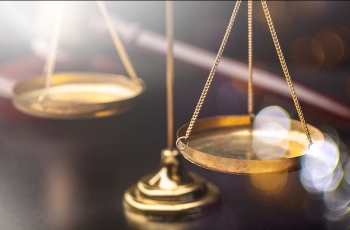 Foto em close de uma balança dourada sobre uma mesa de madeira. A balança é um dos símbolos da Justiça e indica equilíbrio e ponderação na hora de pesar, lado a lado, os argumentos contra e a favor dos acusados.