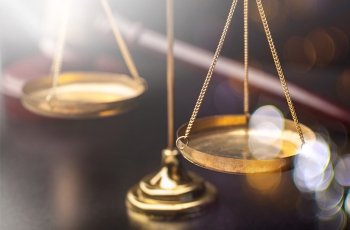 Foto em close de uma balança dourada sobre uma mesa de madeira. A balança é um dos símbolos da Justiça e indica equilíbrio e ponderação na hora de pesar, lado a lado, os argumentos contra e a favor dos acusados.