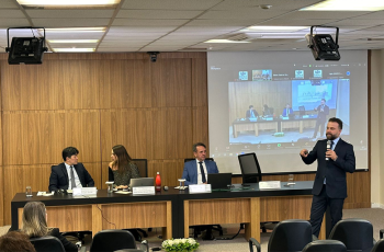 Foto de uma apresentação em um auditório. Um homem branco de cabelo curto, barba e bigode escuro, vestindo terno preto, fala levando um microfone à boca. Atrás dele está uma mesa onde se encontram uma mulher e dois homens sentados e um projetor que transmite uma reunião virtual com enquadramento similar ao da foto.