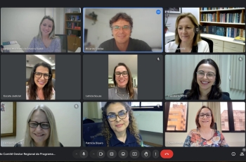 Esta é uma imagem de uma reunião virtual realizada por videoconferência, onde nove pessoas aparecem em suas respectivas janelas de vídeo, organizadas em uma grade. Todas as pessoas estão sorrindo, e há uma variedade de fundos, como estantes de livros e janelas, que mostram diferentes ambientes de trabalho. Os participantes parecem estar concentrados na reunião e estão todos vestidos de maneira profissional.