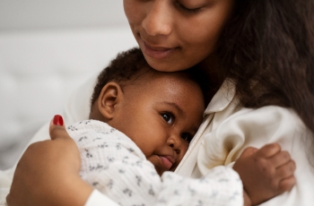 Fotografia ilustrativa de mulher negra com bebê no colo. Ambos vestem roupas claras.
