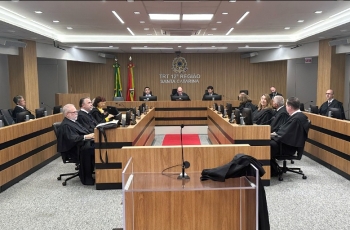 Em um plenário estão sentadas quinze pessoas, sendo três mulheres e doze homens. Eles vestem togas pretas e estão em estações de trabalho posicionadas em quatro fileiras. Ao fundo, estão sentadoes três homens, sendo ao centro, o presidente do TRT-SC.