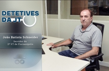 Detetives da JT: João Batista Schneider, servidor da 2ª VT de Florianópolis. Um homem branco sentando à mesa de trabalho, vestindo camisa polo cinza e olhando para a câmera