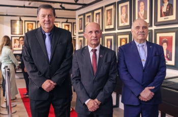 Três homens de terno, em pé, sorriem. Eles estão à frente de uma galeria de quadros