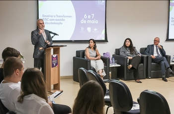 Homem branco, grisalho, de terno cinza e gravata em frente a um púlpito dando uma palestra