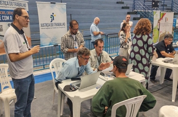 Um homem, trajando um moleton verde escuro e um boné preto está sentado em um posto de atendimento, no qual dois homens fornecem informações. Outros dois homens estão em pé, ao lado da mesa. Ao fundo há dois banners nos quais se lê: Justiça do Trabalho.